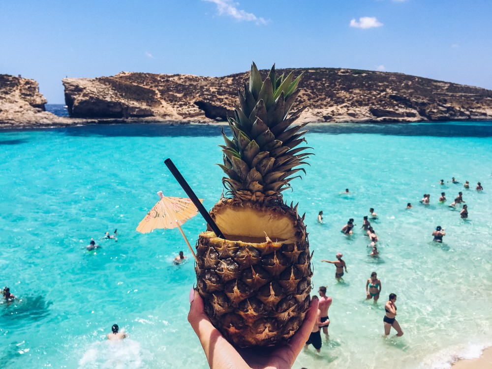 An image of a pineapple cocktail with people swimming and playing in the Blue Lagoon's water in the background.