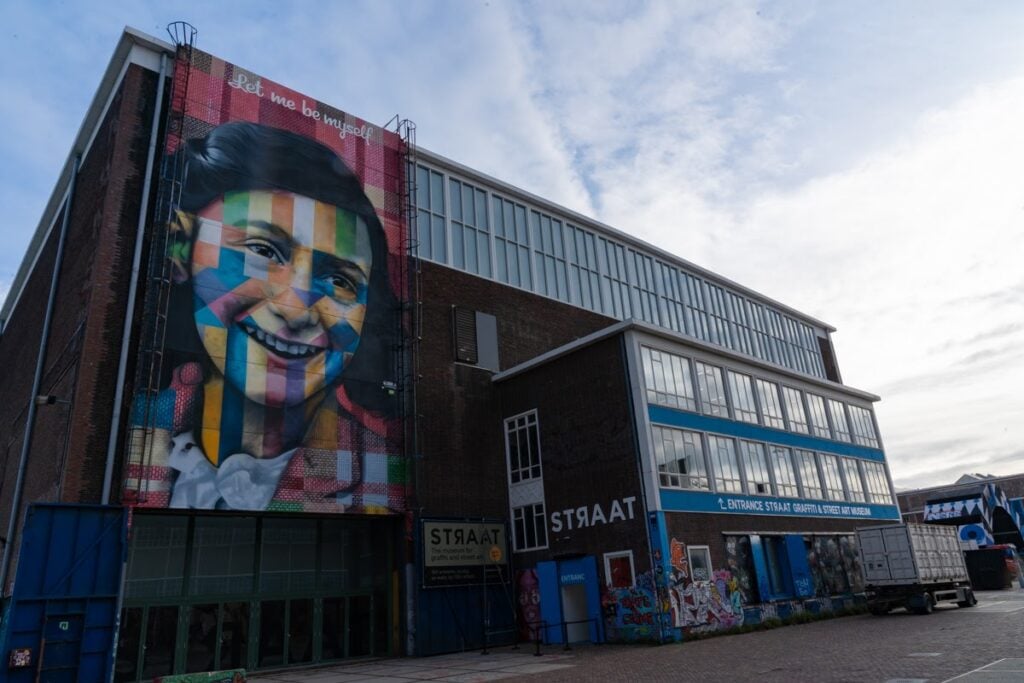The exterior of STRAAT Museum, featuring a colourful mural of Anne Frank and various tags and graffiti.