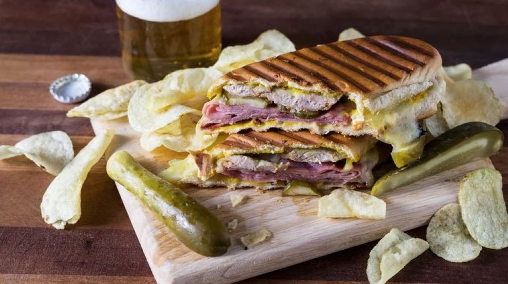 A Cuban sandwich accompanied by chips, pickles, and a beer, a unique thing to try in Tampa